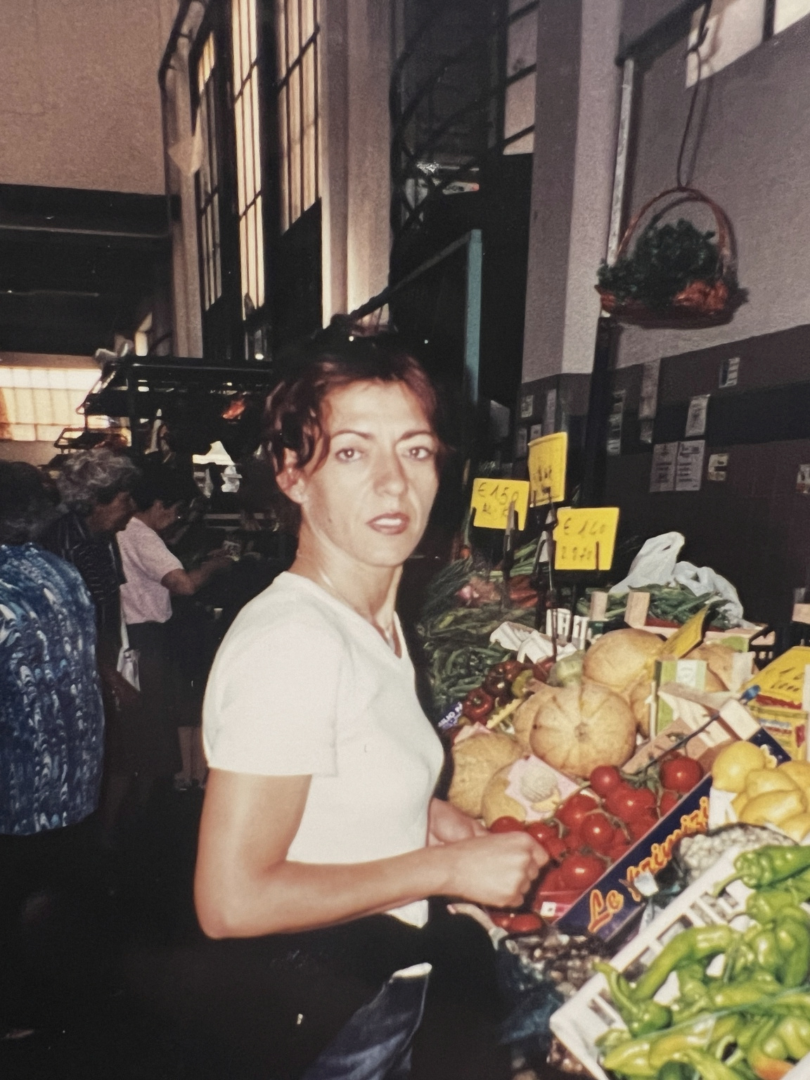 A woman at an Italian market stall beside crates of tomatoes and peppers