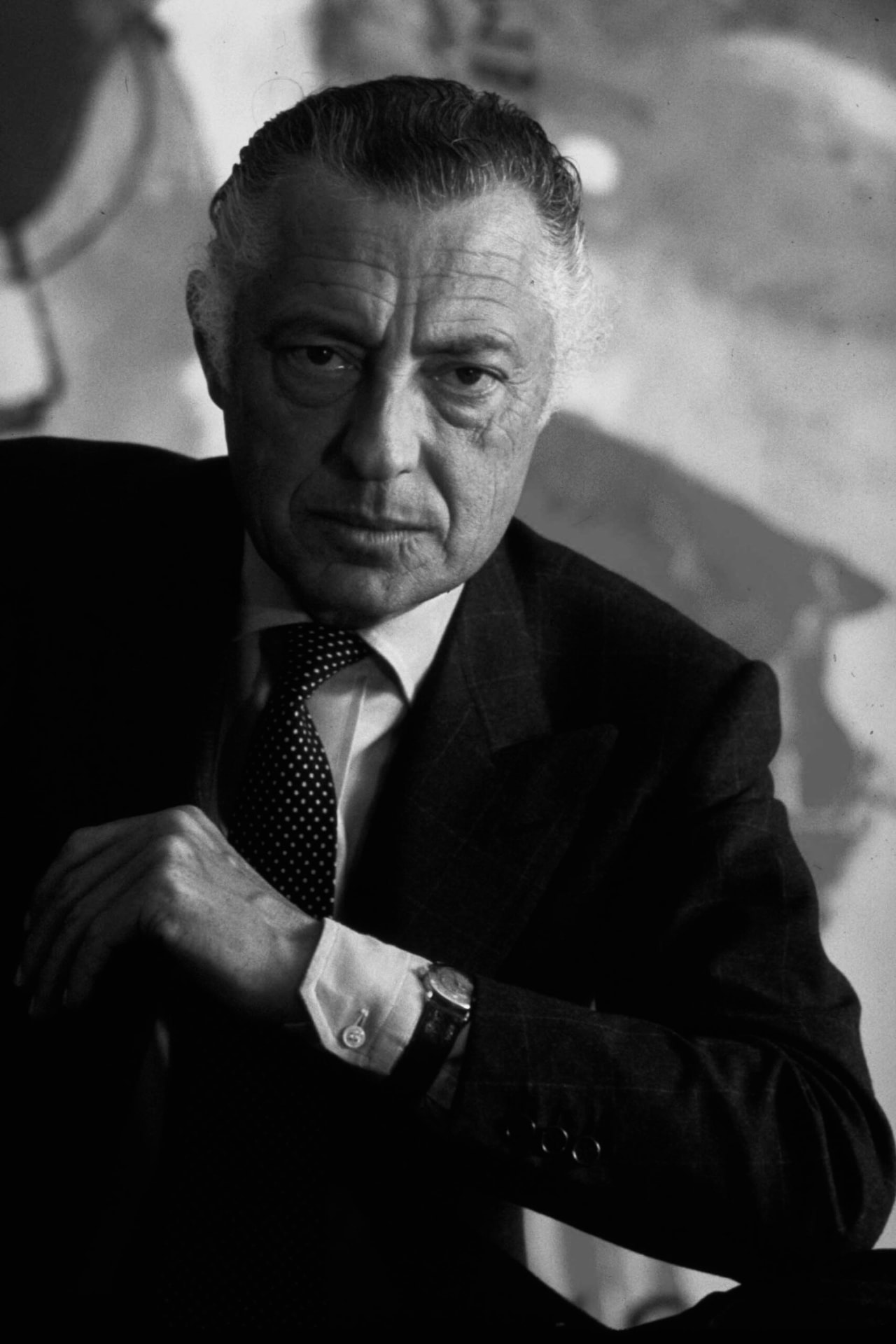 A black and white portrait of Gianni Agnelli wearing a watch over his shirt cuff, in a windowpane-check suit and tie