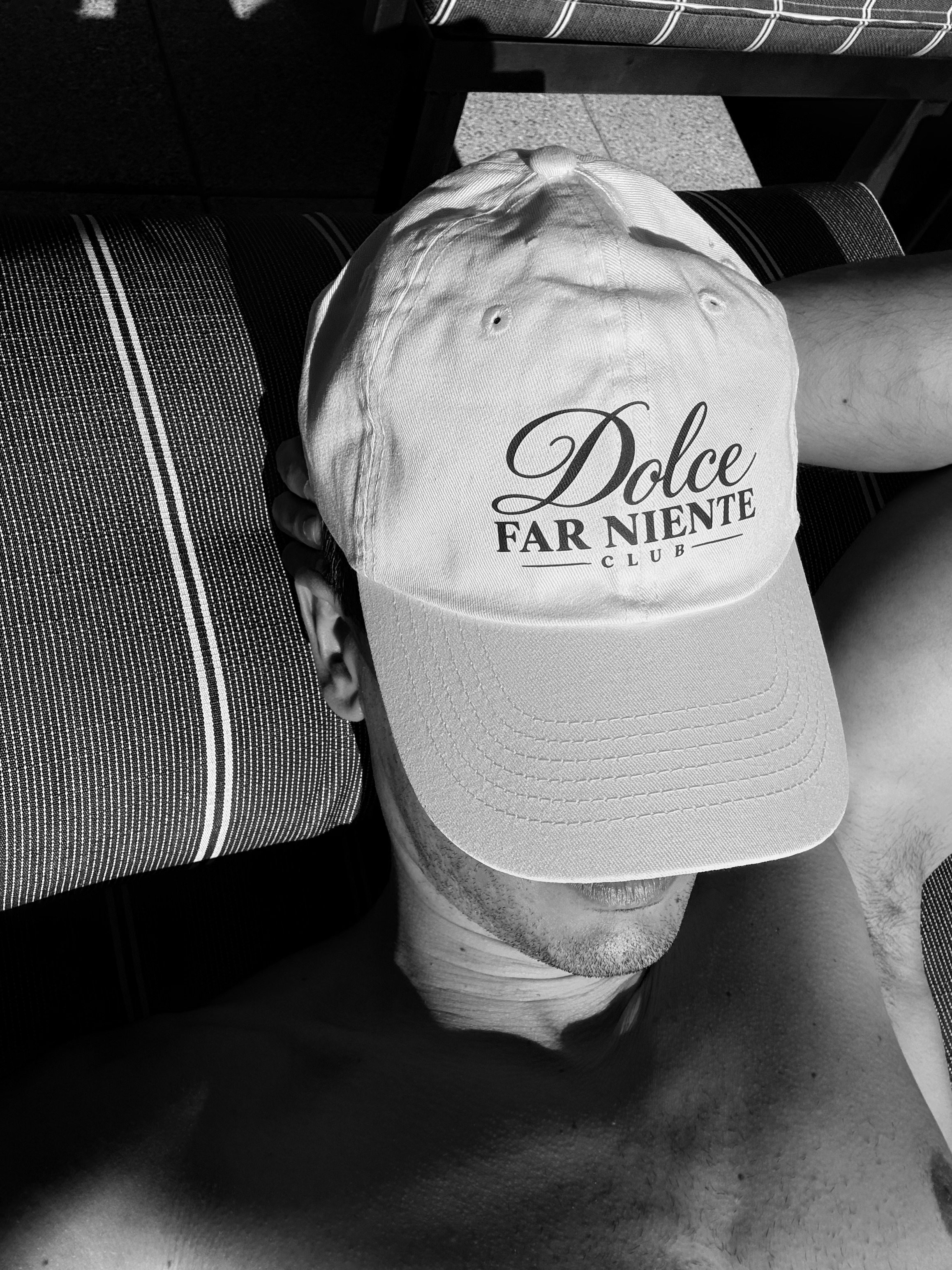 A black and white photograph of a man asleep on a striped lounger, a Dolce Far Niente Club cap over his face