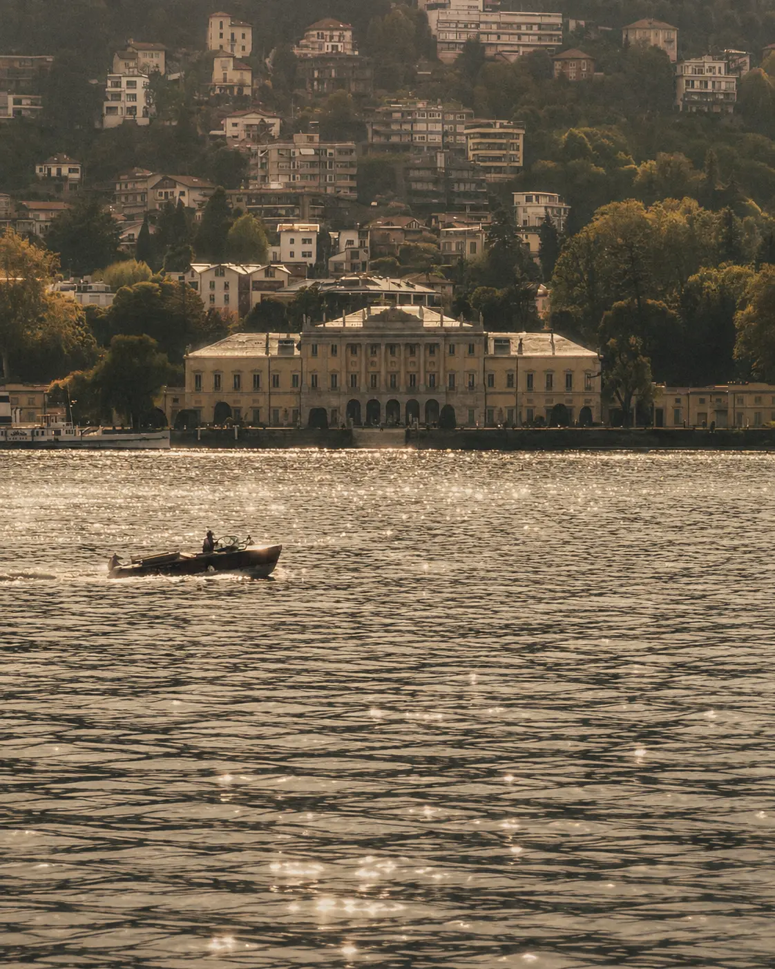 A small boat crossing sunlit water below a wooded lakeside town