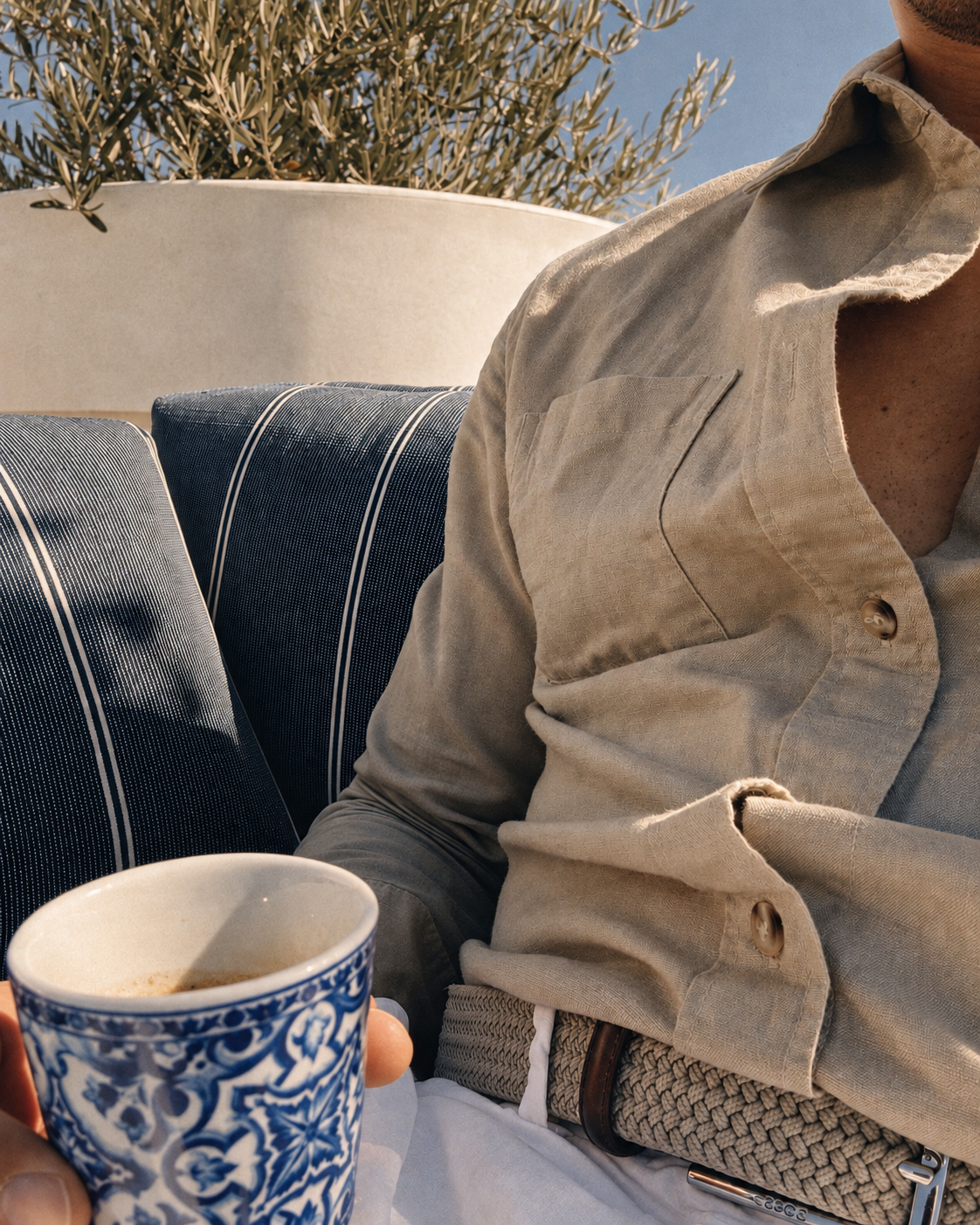 A person in an unbuttoned linen shirt holding a blue and white patterned coffee cup, seated on a navy striped bench beside an olive tree in the sun