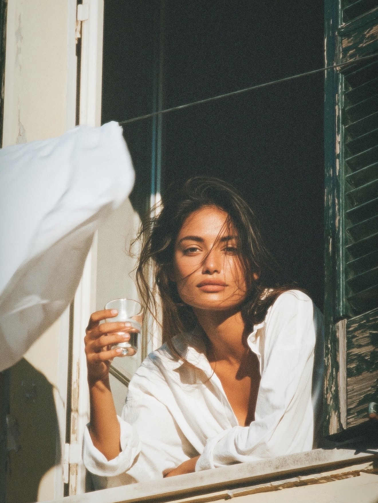 A woman leaning from a shuttered window in a white shirt, holding a glass of water